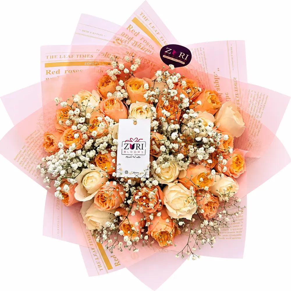 Bouquet of orange and beige roses with baby's breath on pink paper, featuring Zuri Flowers brand.